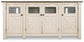 The Bolanburg Extra Large TV Stand is a white wooden sideboard with four doors featuring X-patterns and glass panels, dark handles, and a contrasting dark top for a stylish two-tone farmhouse look.