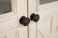 Close-up of two black, octagonal door knobs on white, rustic wooden cabinet doors—perfect farmhouse-style details found on the Bolanburg Extra Large TV Stand.