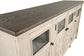 The Bolanburg Extra Large TV Stand features a light wood frame and dark wood top, glass-front cabinet doors, and black handles, combining rustic farmhouse charm with modern style for your living space.