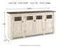 The Bolanburg Extra Large TV Stand features a white and brown two-tone finish, four doors with glass panels, measures 36"H x 74.25"W x 18"D, weighs 162 lbs, and is ideal for a farmhouse-inspired living room.