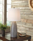 The Shavontae Poly Table Lamp (2/CN) with a white shade sits on a wooden table beside a potted plant and glass box, set against a wood-paneled wall.