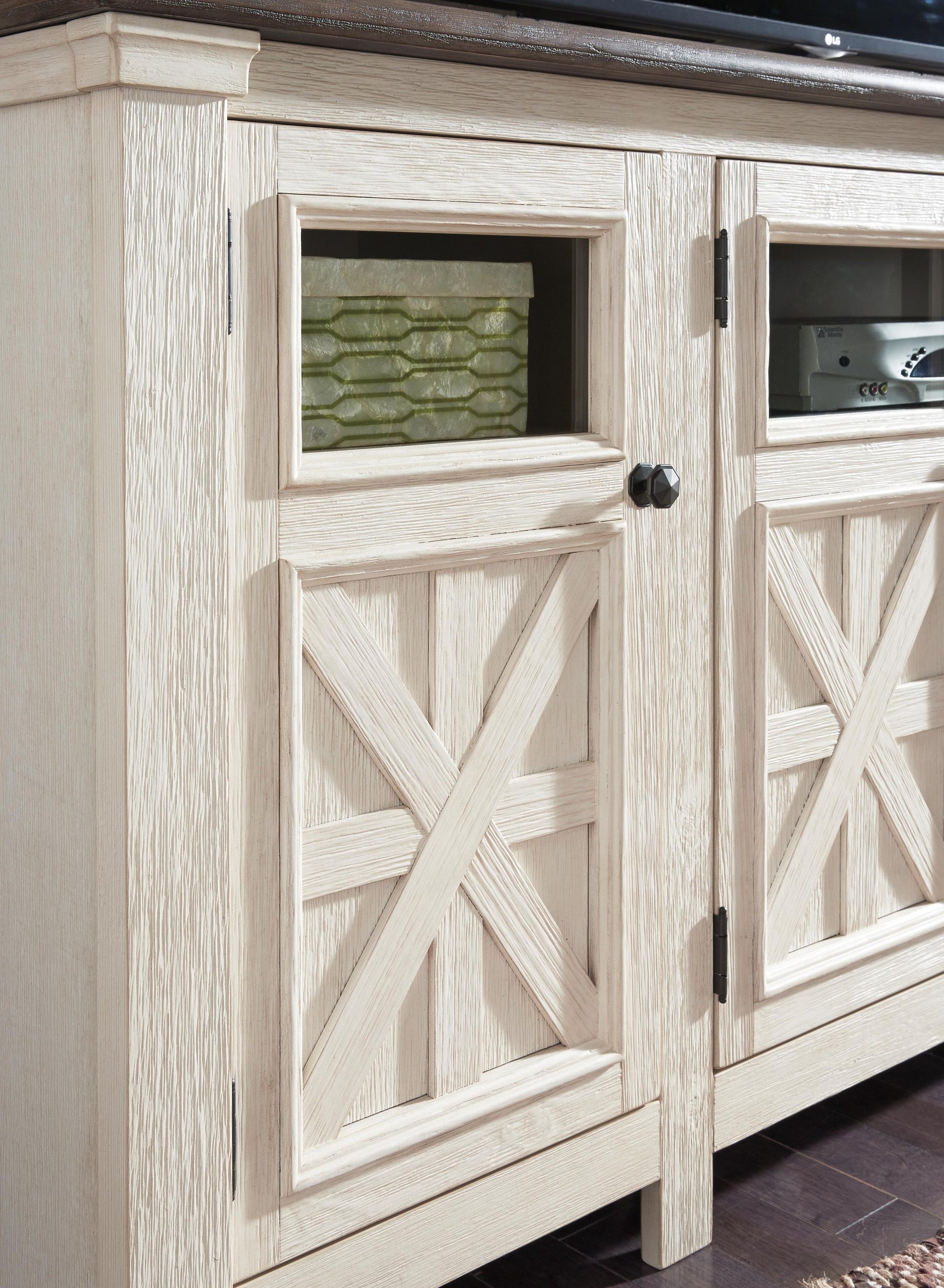 A close-up of the Bolanburg Extra Large TV Stand showcases its light wood finish, decorative crossbuck doors, and a glass-fronted upper section displaying a green object.