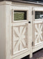 A close-up of the Bolanburg Extra Large TV Stand showcases its light wood finish, decorative crossbuck doors, and a glass-fronted upper section displaying a green object.