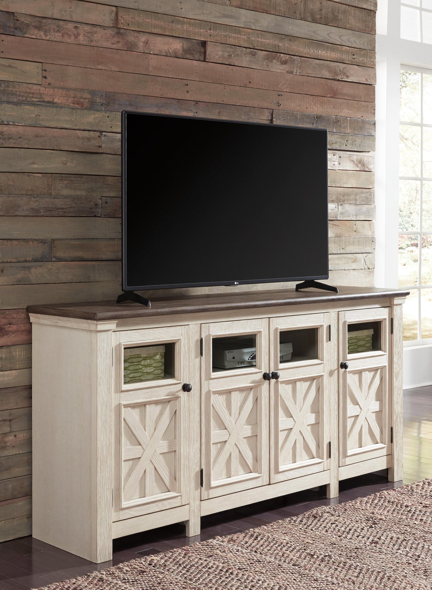 A flat-screen TV rests on the Bolanburg Extra Large TV Stand, featuring four cabinet doors with glass panels, positioned against a rustic wood plank wall in a bright room.