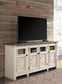 A flat-screen TV rests on the Bolanburg Extra Large TV Stand, featuring four cabinet doors with glass panels, positioned against a rustic wood plank wall in a bright room.