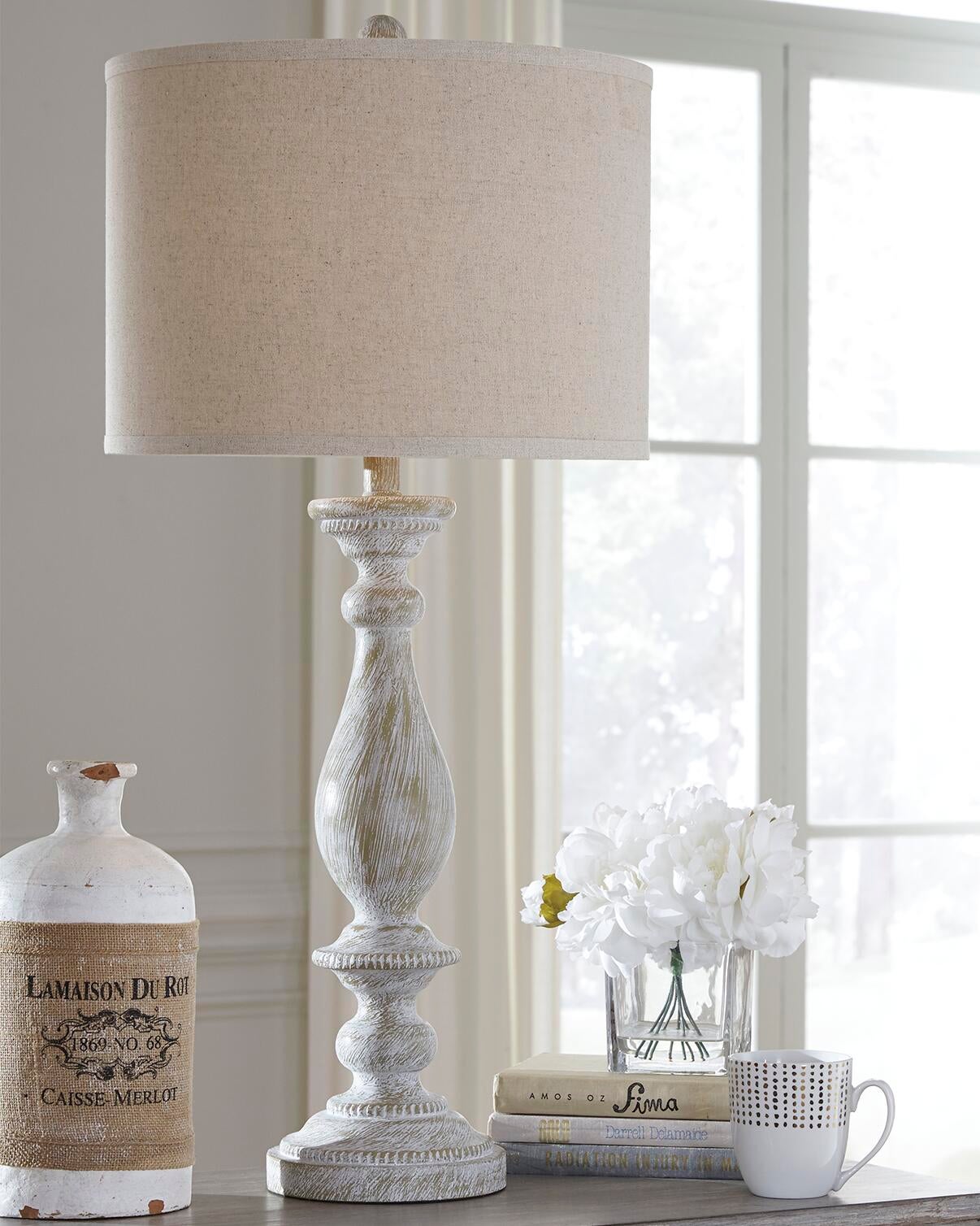 A Bernadate Poly Table Lamp, a ceramic jug, a stack of books, a white mug, and a vase with white flowers sit on a table by the window.