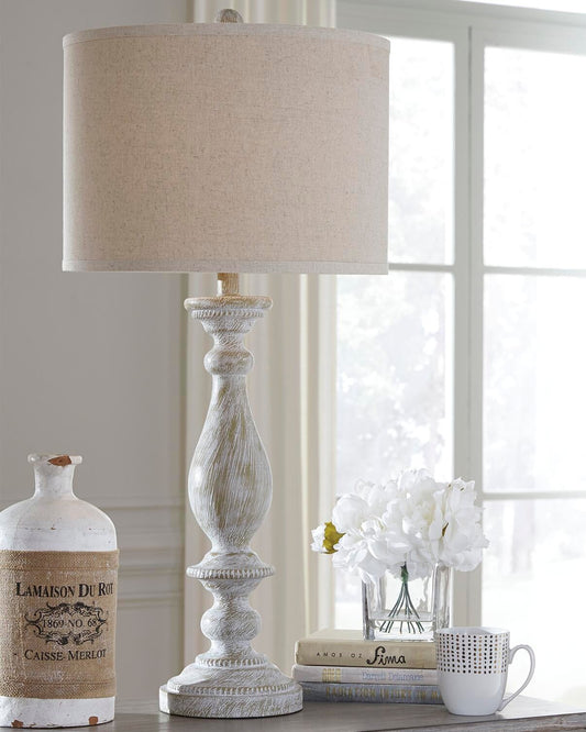A Bernadate Poly Table Lamp, a ceramic jug, a stack of books, a white mug, and a vase with white flowers sit on a table by the window.