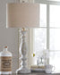 A Bernadate Poly Table Lamp, a ceramic jug, a stack of books, a white mug, and a vase with white flowers sit on a table by the window.