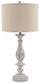 The Bernadate Poly Table Lamp (2/CN) features a beige drum shade, a textured base with a distressed whitewash finish, and ornate candlestick-style detailing.