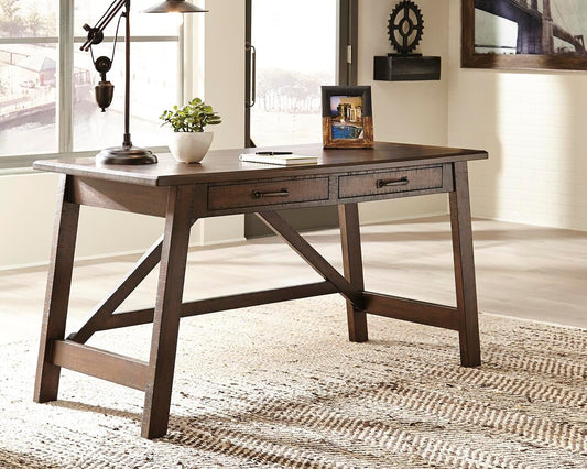 The Baldridge Home Office Large Leg Desk with two drawers stands on a woven rug, holding a lamp, small plant, and framed photo in a bright room with large windows.