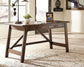 The Baldridge Home Office Large Leg Desk with two drawers stands on a woven rug, holding a lamp, small plant, and framed photo in a bright room with large windows.