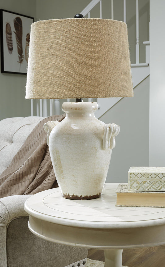 The Emelda Ceramic Table Lamp with a beige fabric shade sits on a round white side table by the sofa and stacked books, with a staircase and framed art in the background.