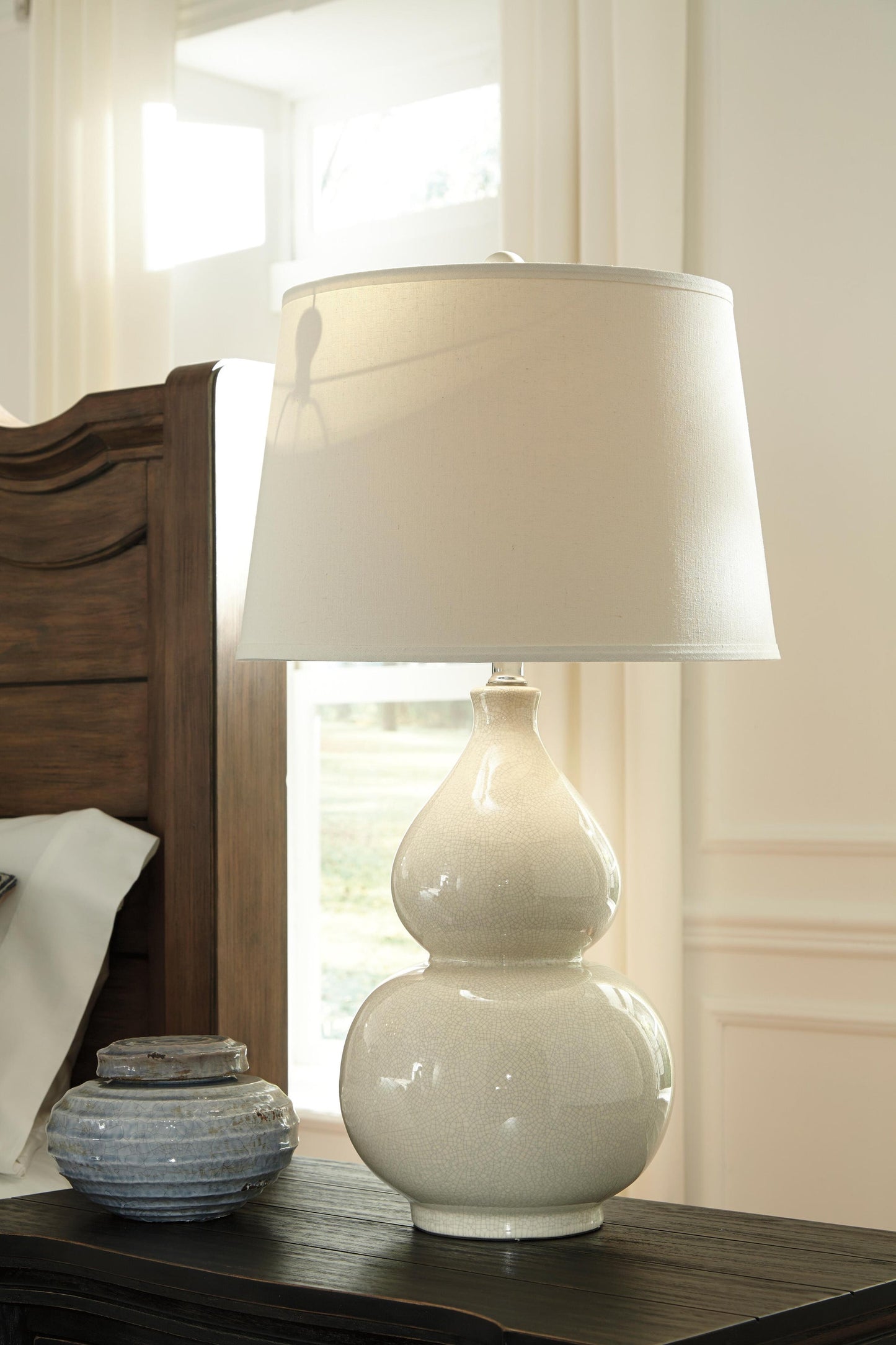 The Saffi Ceramic Table Lamp (1/CN) with a fabric shade sits on a dark wood nightstand by a wooden headboard and decorative jar, providing stylish contemporary lighting.