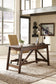 A modern office features the Baldridge Home Office Large Leg Desk, a rolling chair, desk lamp, potted plant, wall art, large windows, and rustic industrial decor.