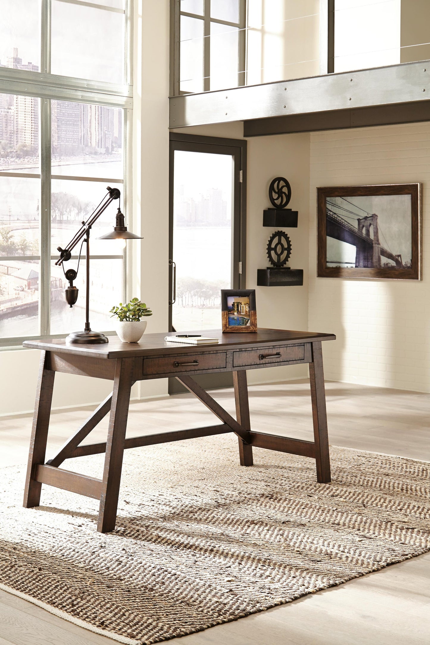 The Baldridge Home Office Large Leg Desk, adorned with a lamp, plant, and photo frame, sits on a woven rug in a bright office featuring large windows with cityscape views.