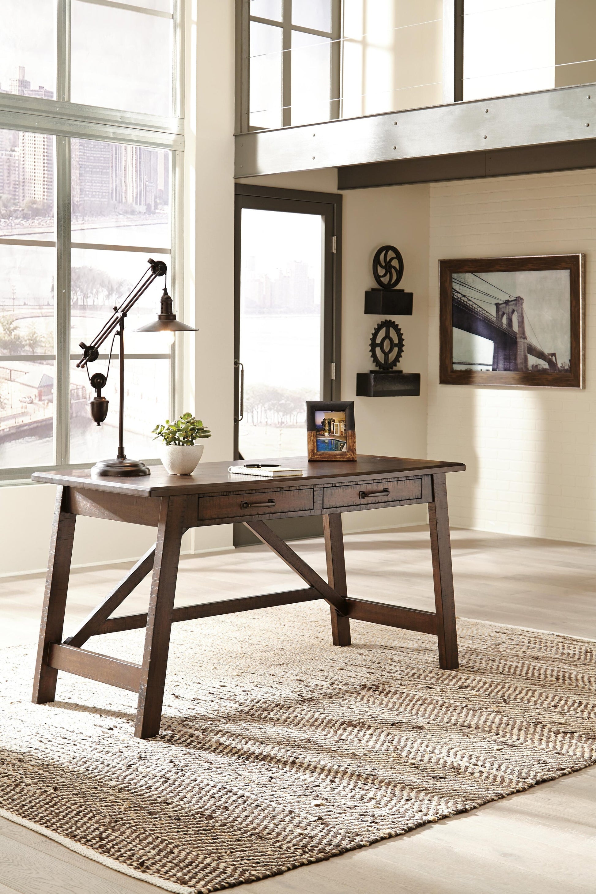 The Baldridge Home Office Large Leg Desk, adorned with a lamp, plant, and photo frame, sits on a woven rug in a bright office featuring large windows with cityscape views.