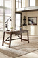 The Baldridge Home Office Large Leg Desk, adorned with a lamp, plant, and photo frame, sits on a woven rug in a bright office featuring large windows with cityscape views.
