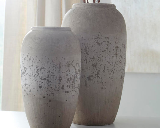 The Dimitra Vase Set (2/CN) features two tall, weathered ceramic vases with a distressed gray finish, creating vintage-inspired decorative accents on a white surface in front of sheer curtains.