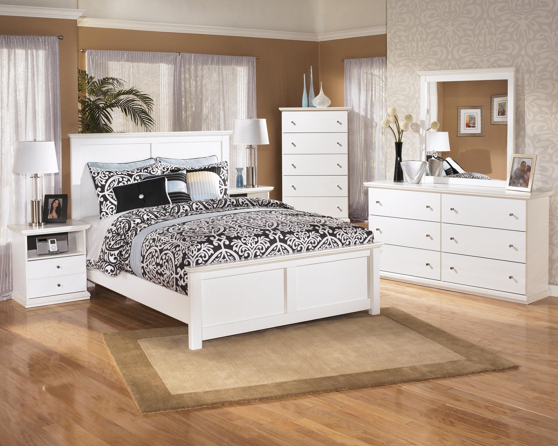 A bedroom with white furniture, including a bed, nightstands, cottage-chic dresser with mirror, the Bostwick Shoals Five Drawer Chest, and patterned bedding on a rug over hardwood floors.