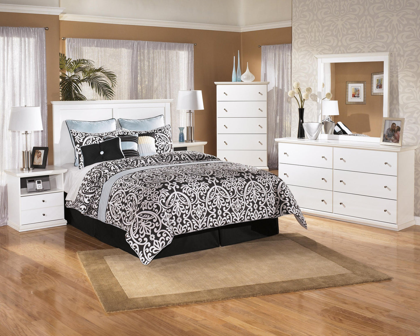 A neatly arranged bedroom features a white bed, Bostwick Shoals Five Drawer Chest, nightstands, lamps, and patterned bedding on a beige rug over wood flooring.