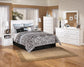 A neatly arranged bedroom features a white bed, Bostwick Shoals Five Drawer Chest, nightstands, lamps, and patterned bedding on a beige rug over wood flooring.