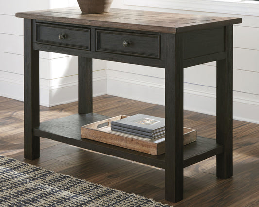 The Tyler Creek Sofa Table features two drawers with traditional hardware, crafted from weathered gray-brown wood. It stands on a wood floor by a window, with a tray of books on the lower shelf and a vase placed on top.