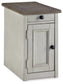 The Bolanburg Chair Side End Table is a small, rectangular table in antique white with a single drawer, cabinet door, black round knobs, and built-in USB ports.