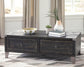 The Todoe Lift Top Cocktail Table, featuring dark wood and two drawers, is styled with a bowl, potted plant, vase with greenery, and two stacked books on a patterned rug for a rustic-industrial vibe.