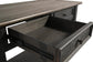 A close-up of the Tyler Creek Sofa Table shows its weathered gray-brown wood finish and an open dark wood drawer, highlighting traditional hardware on the metal slides.