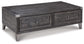 The Todoe Lift Top Cocktail Table features a rectangular design with a dark finish, metal corner accents, two drawers, and angled legs—blending rustic-industrial style and function in one striking piece.