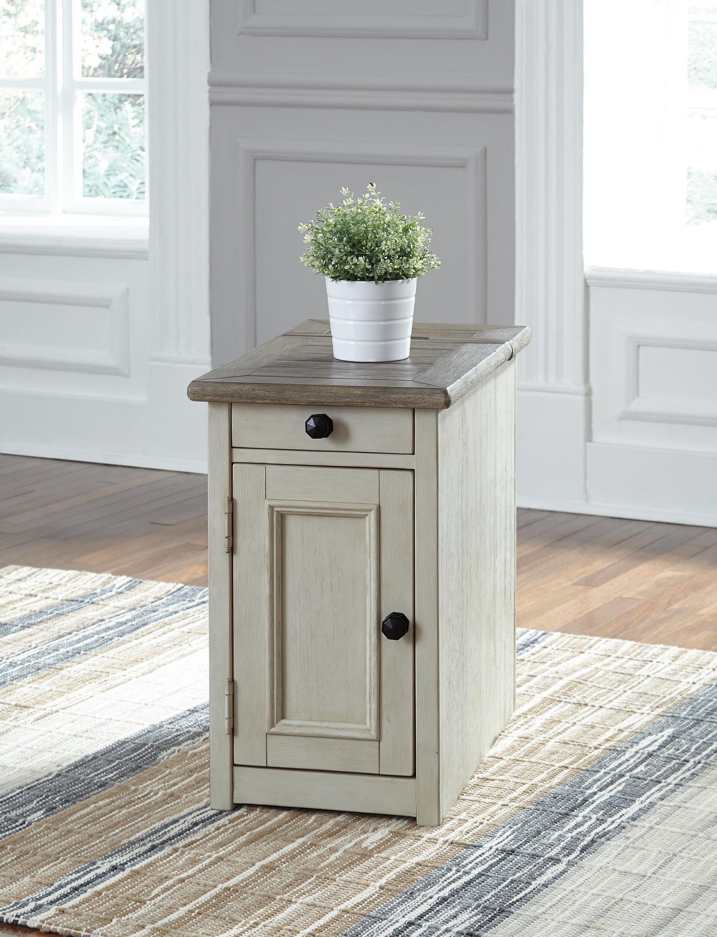 The Bolanburg Chair Side End Table, featuring an antique white finish, drawer, and cabinet door, sits atop a striped rug in a bright room with white paneled walls and is topped with a potted plant.