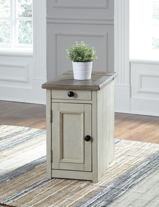 The Bolanburg Chair Side End Table, featuring an antique white finish, drawer, and cabinet door, sits atop a striped rug in a bright room with white paneled walls and is topped with a potted plant.