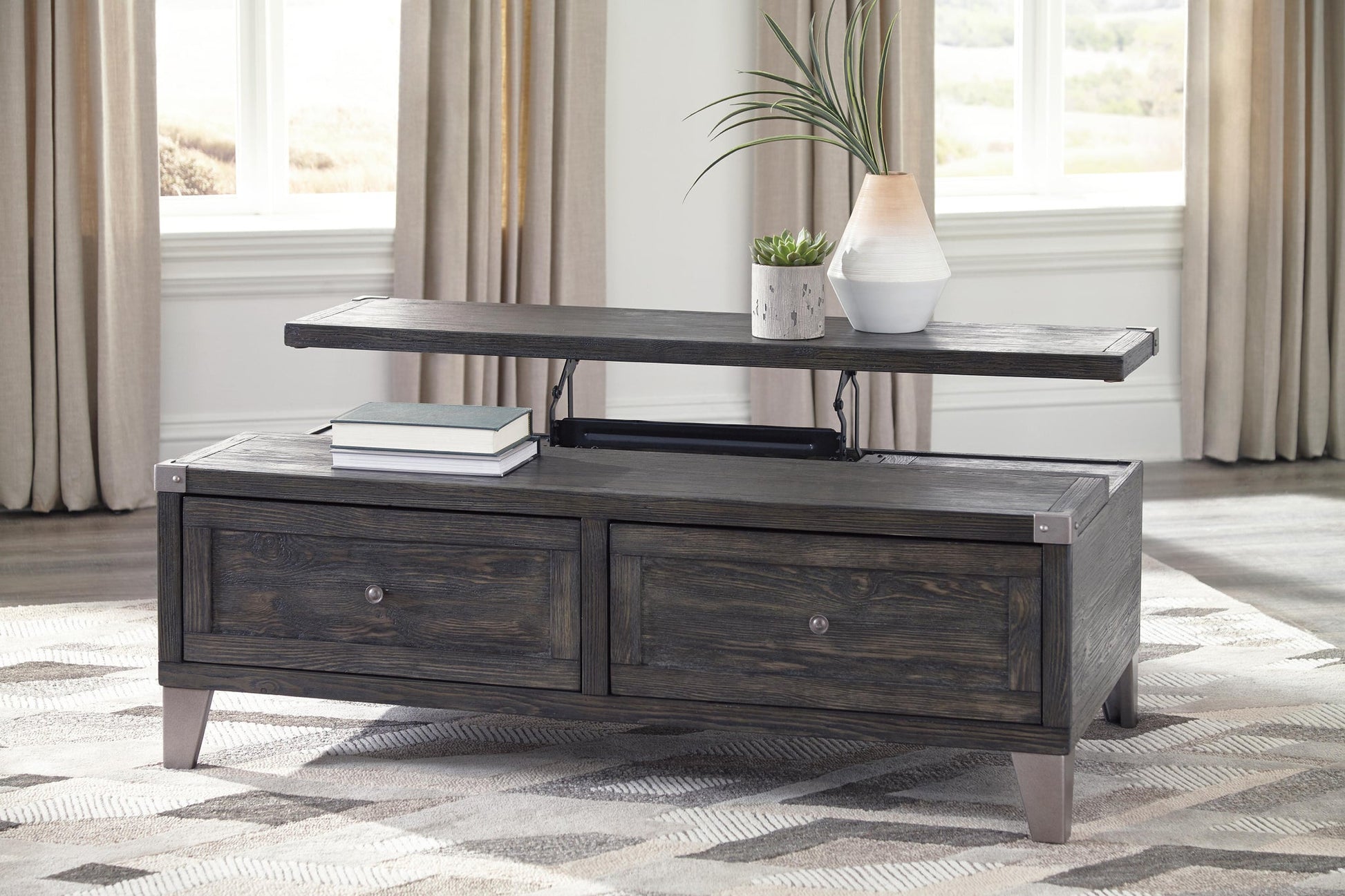 The Todoe Lift Top Cocktail Table, featuring a dark wood finish and two drawers, displays stacked books, a potted plant, and a vase. It's set in a bright room with large windows and beige curtains for a rustic-industrial look.