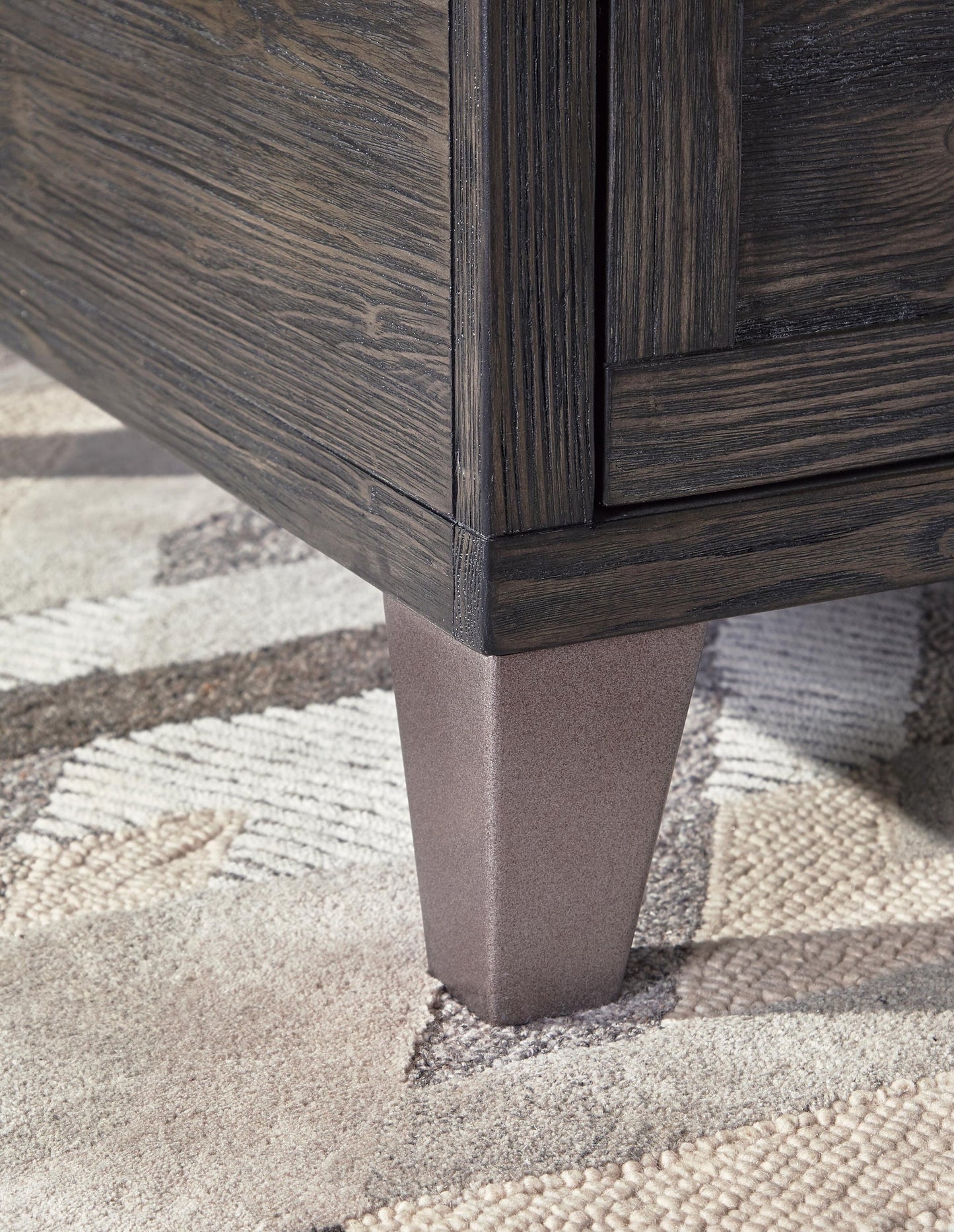 Close-up of the bottom corner of the Todoe Lift Top Cocktail Table, featuring a dark wood finish and tapered metallic leg, set on a neutral-toned textured rug.