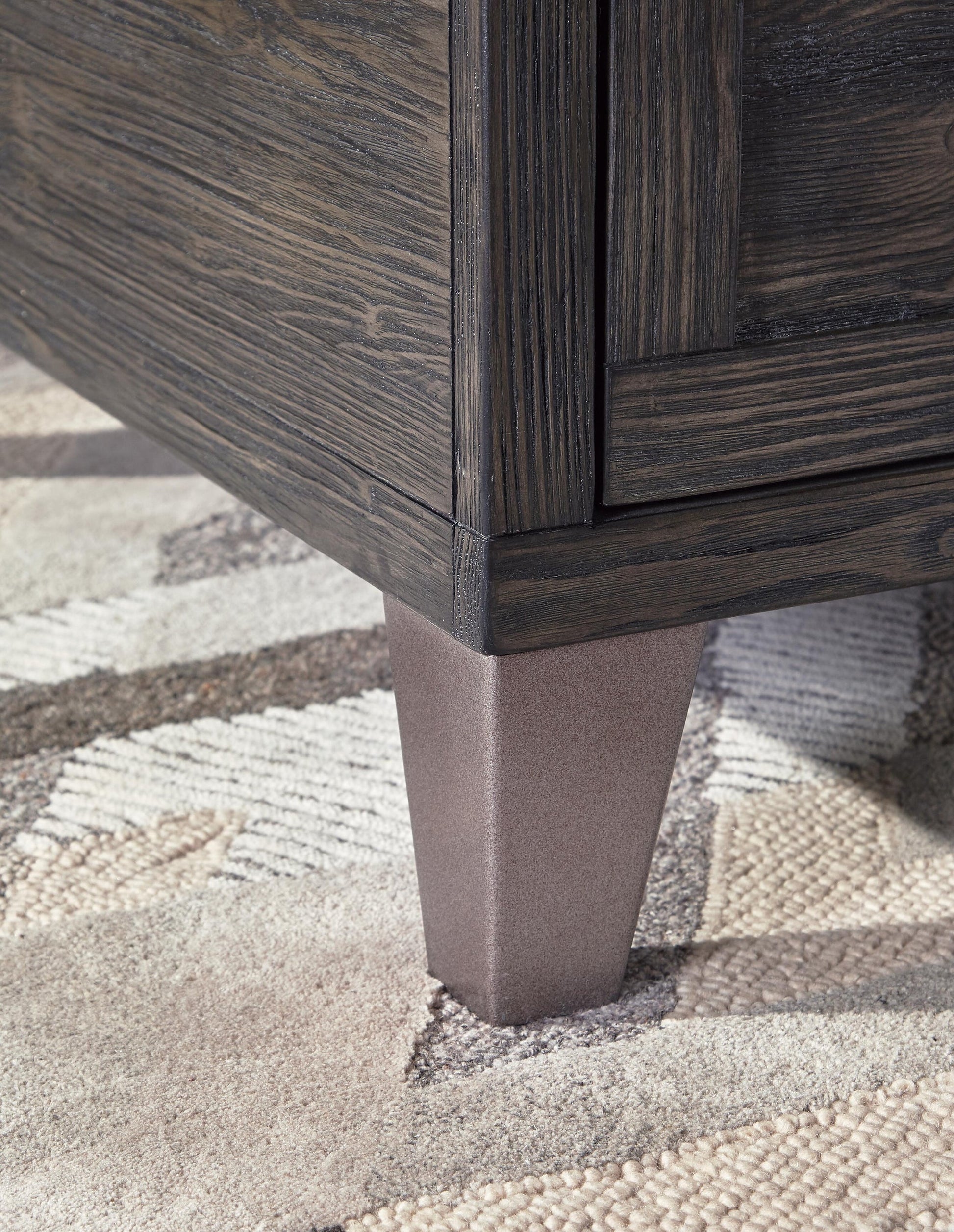 Close-up of the bottom corner of the Todoe Lift Top Cocktail Table, featuring a dark wood finish and tapered metallic leg, set on a neutral-toned textured rug.