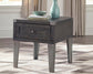 The Todoe Rectangular End Table features a dark wood drawer, metal legs, and corner accents. Topped with a vase, bowl, and books on a patterned rug in a bright room, it stylishly blends rustic-industrial charm with function.
