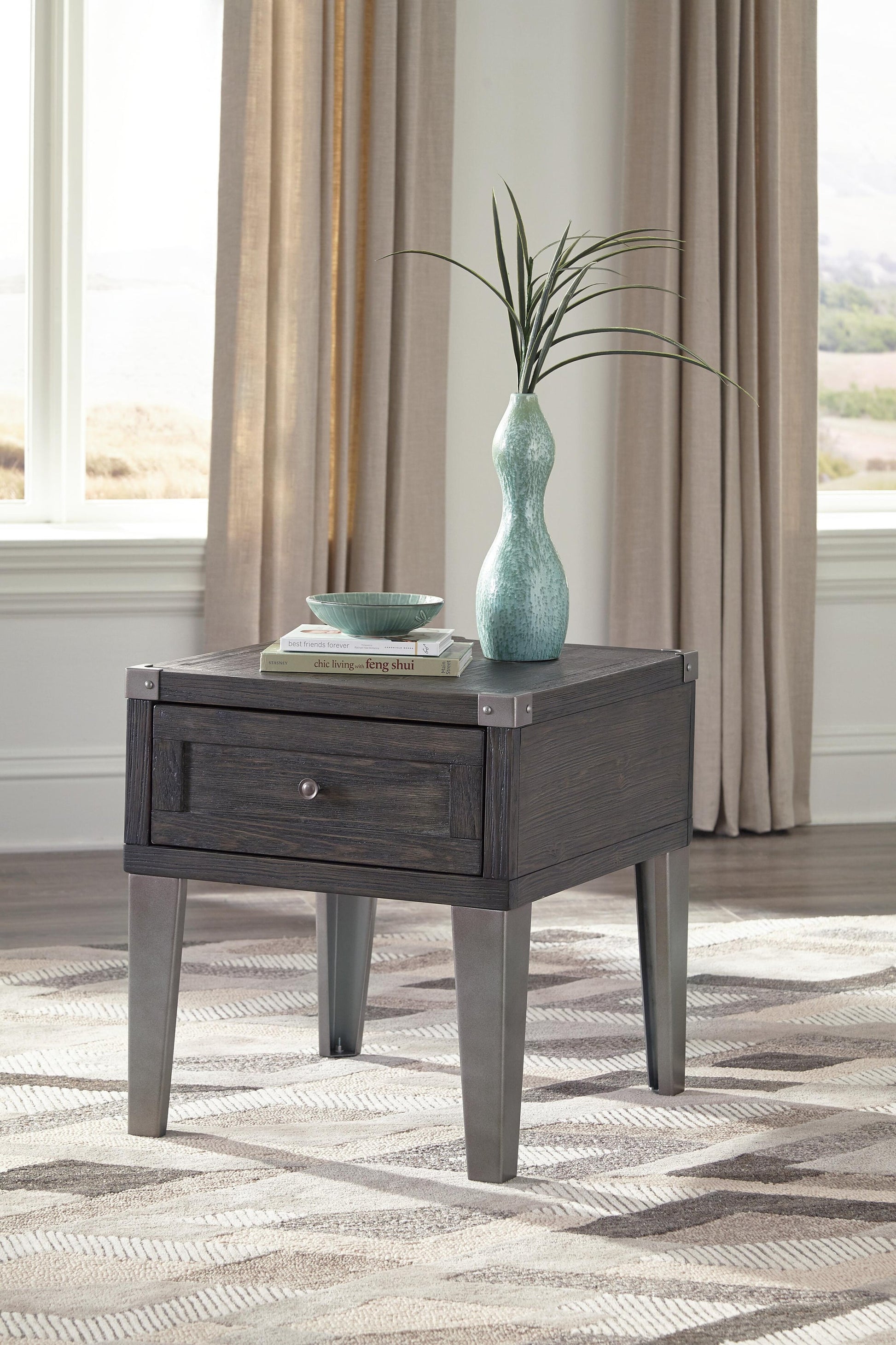 The Todoe Rectangular End Table with USB charging ports holds a drawer, a turquoise vase with greenery, a bowl, and two stacked books. Beige curtains and a window serve as the backdrop for this stylish piece.