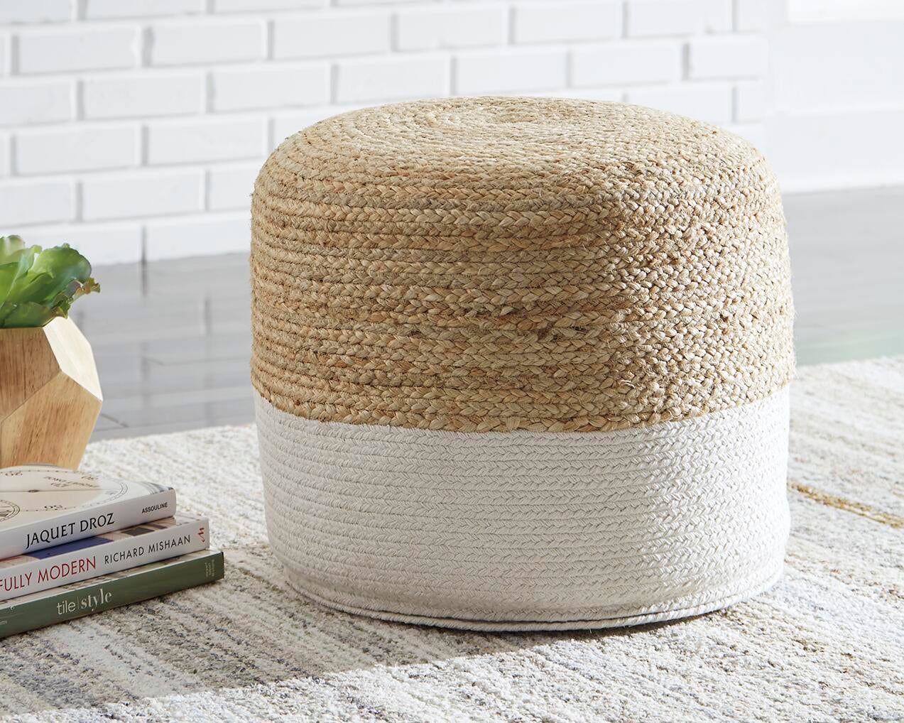 The Sweed Valley Pouf, featuring a round jute design with a natural top and white bottom, sits on a patterned rug beside books and a small plant on a wooden stand.