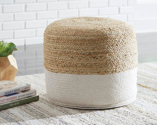 The Sweed Valley Pouf, featuring a round jute design with a natural top and white bottom, sits on a patterned rug beside books and a small plant on a wooden stand.
