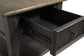 A close-up of the Tyler Creek Rectangular End Table shows its weathered gray-brown top and an open drawer revealing the interior and metal sliding mechanism.