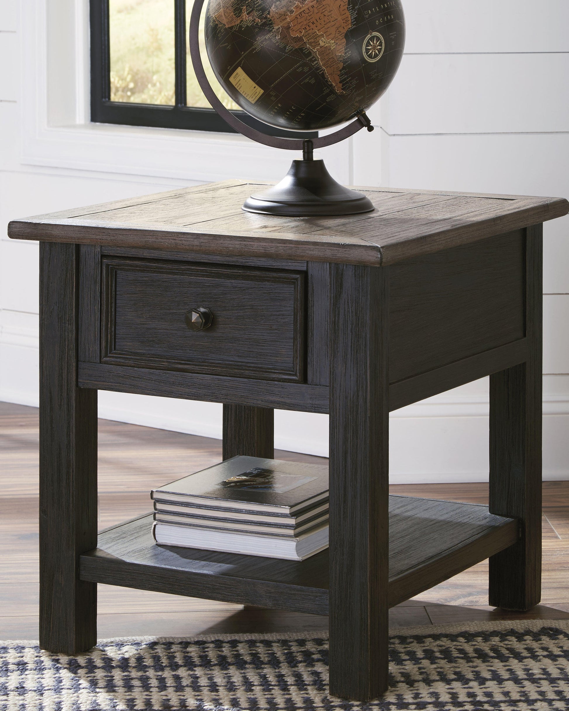 The Tyler Creek Rectangular End Table, with a weathered gray-brown top, includes a drawer and lower shelf. It displays a decorative globe above and stacked books below, set against a white wall and window.