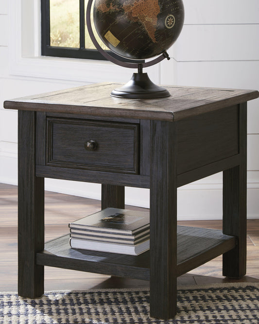 The Tyler Creek Rectangular End Table, with a weathered gray-brown top, includes a drawer and lower shelf. It displays a decorative globe above and stacked books below, set against a white wall and window.
