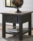 The Tyler Creek Rectangular End Table, with a weathered gray-brown top, includes a drawer and lower shelf. It displays a decorative globe above and stacked books below, set against a white wall and window.