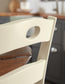 Close-up of the cream-colored Woodanville Dining Room Side Chair (2/CN) featuring a rounded cutout, placed in a kitchen with dark cabinets and a wooden countertop in the background.