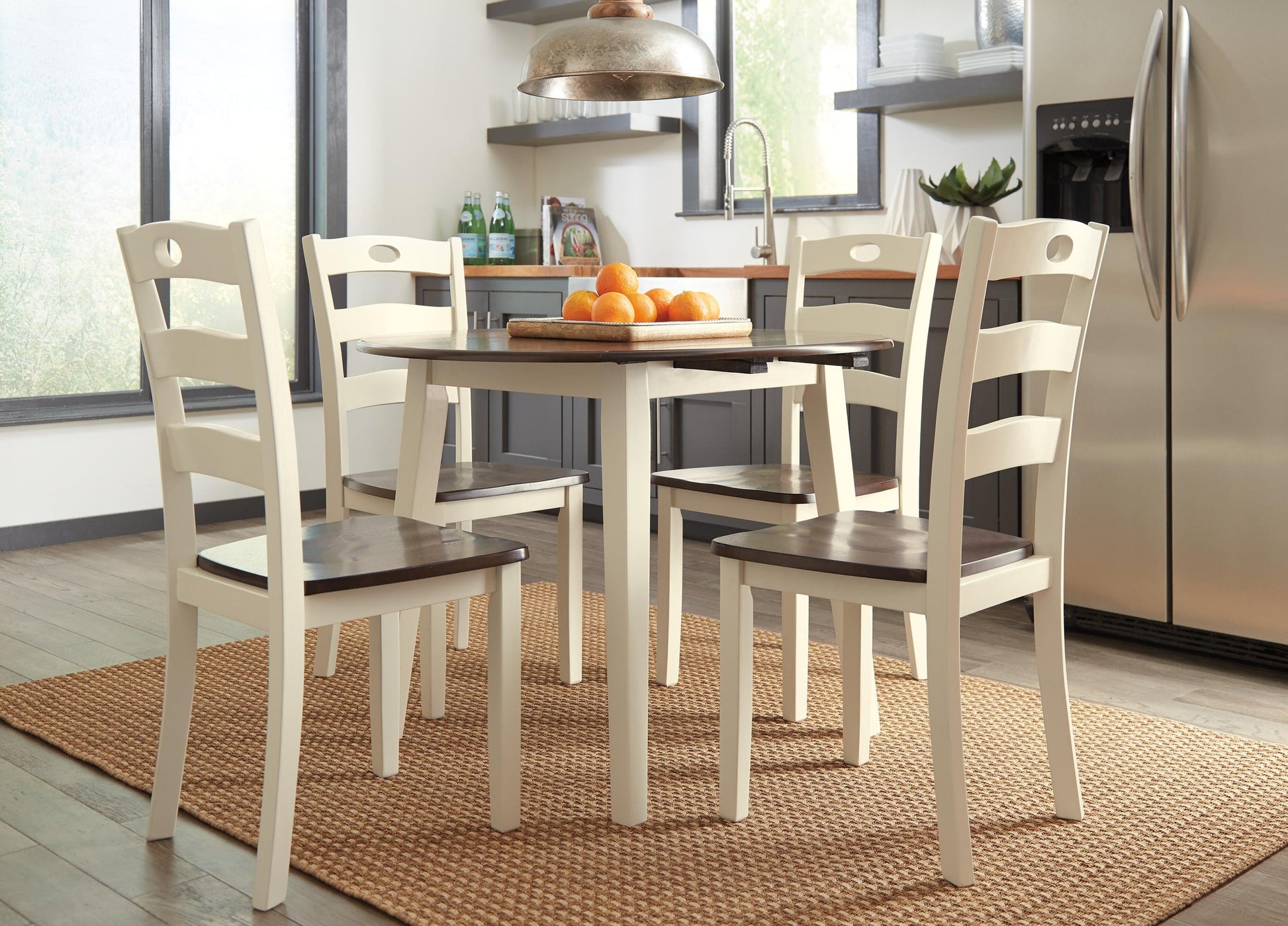 A modern kitchen features a round table with four Woodanville Dining Room Side Chairs, a fruit bowl with oranges, stainless steel appliances, and large windows letting in natural light.