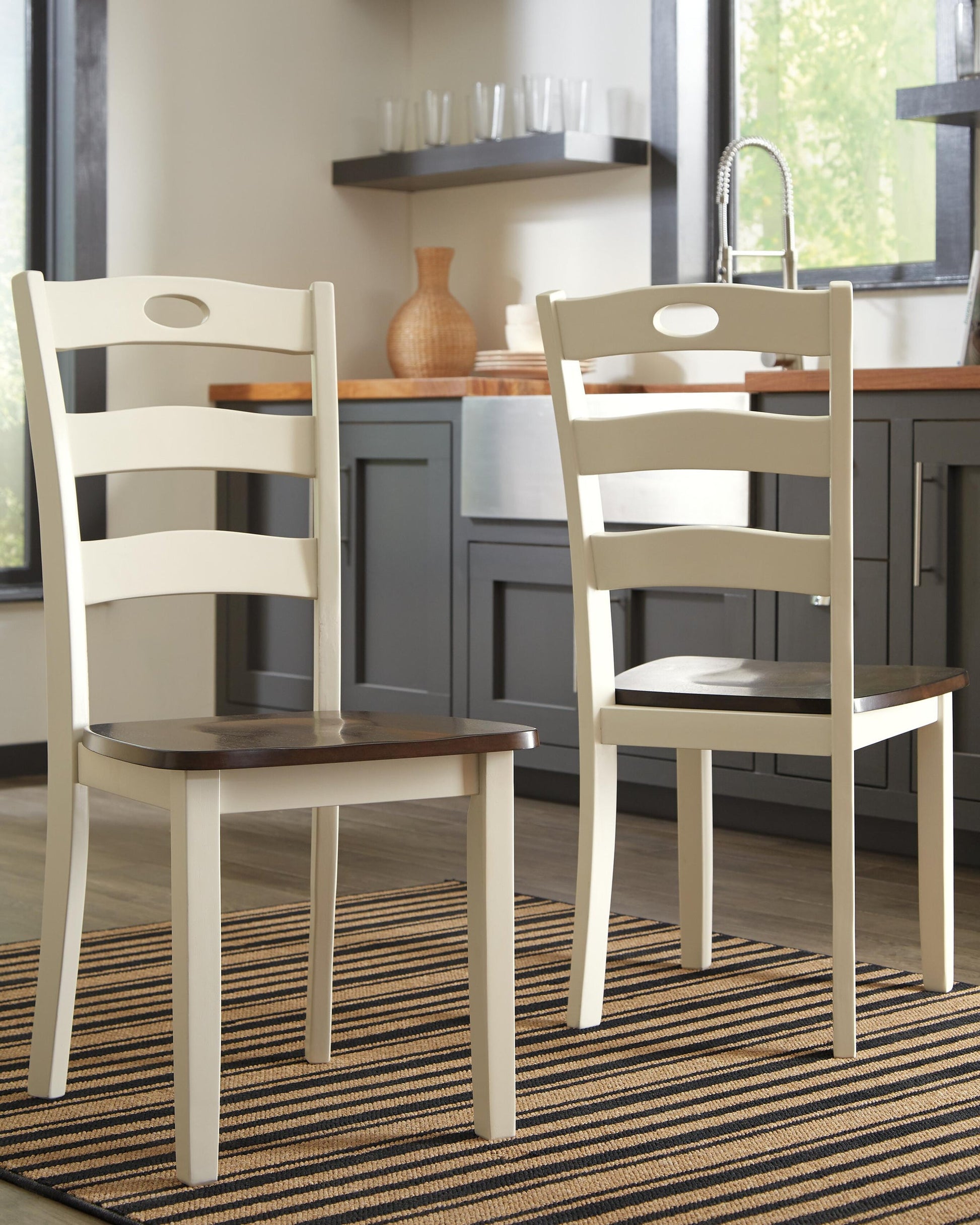 Two Woodanville Dining Room Side Chairs with cream frames and dark brown seats sit on a striped rug in a kitchen featuring gray cabinets and a window.