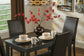 The Kimonte Rectangular Dining Room Table, paired with four black chairs, is set with plates, bowls, mugs, and features a dark vase with red flowers as a centerpiece under a geometric pendant light.