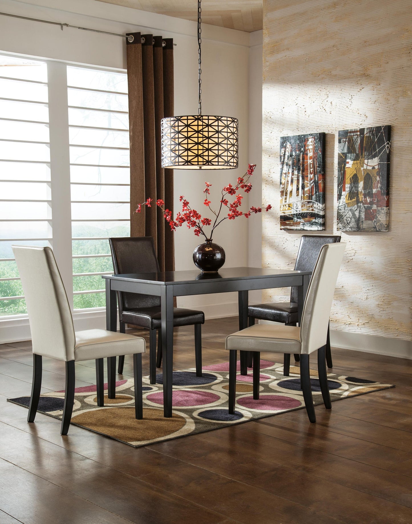 A small dining area featuring the Kimonte Rectangular Dining Room Table, four chairs, a vase with red flowers, a patterned rug, hanging light fixture, two wall artworks, and large windows covered with blinds.