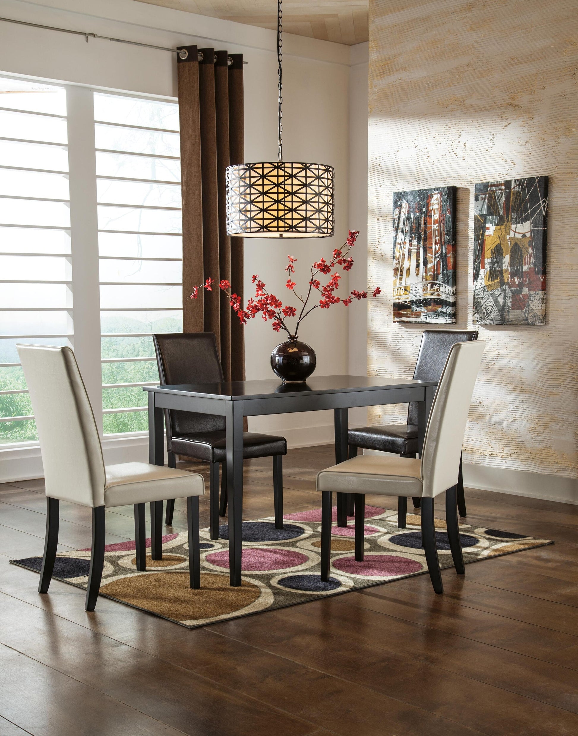 A small dining area featuring the Kimonte Rectangular Dining Room Table, four chairs, a vase with red flowers, a patterned rug, hanging light fixture, two wall artworks, and large windows covered with blinds.