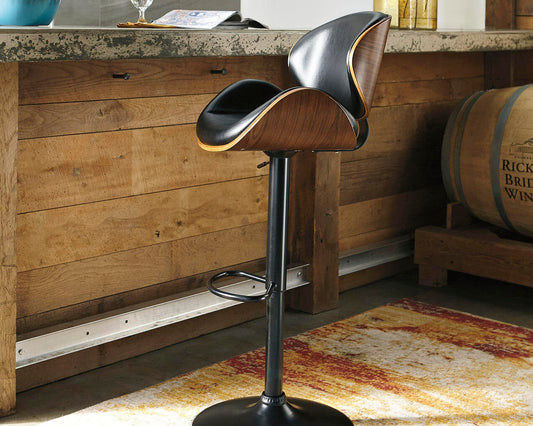 The Bellatier Tall UPH Swivel Barstool features adjustable height, a black metal base, wood accents, and a black faux leather cushioned seat, shown at a rustic wood bar counter with a wine barrel beside it.